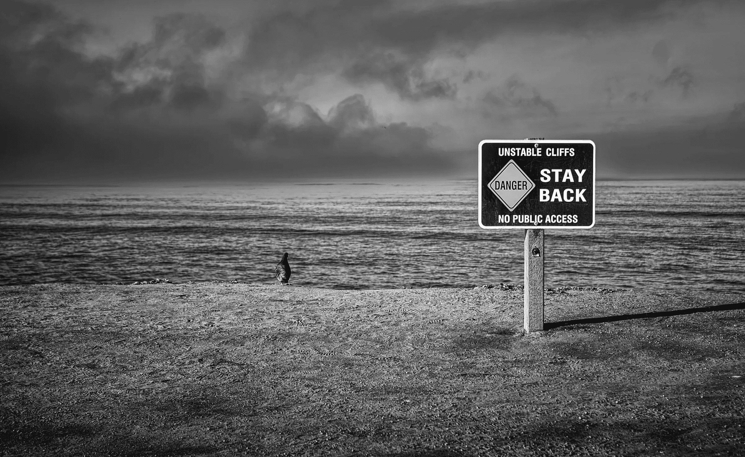Black and white photograph of a warning sign reading 'Unstable Cliffs, Stay Back' on a beach, with the sea and a dramatic cloudy sky in the background.