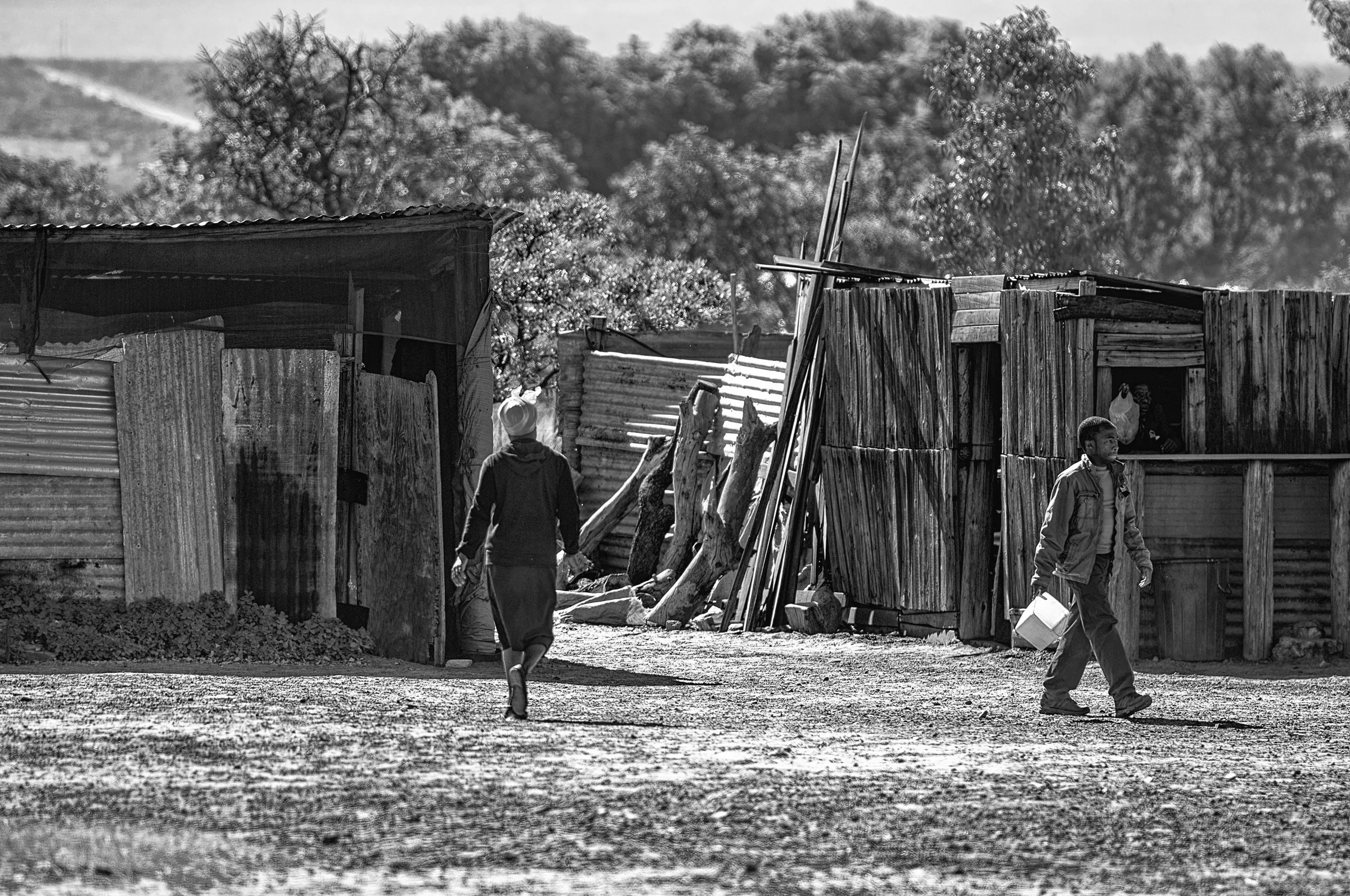 Two people walking between simple dwellings made of corrugated metal and wood in Vaalwater, South Africa.
